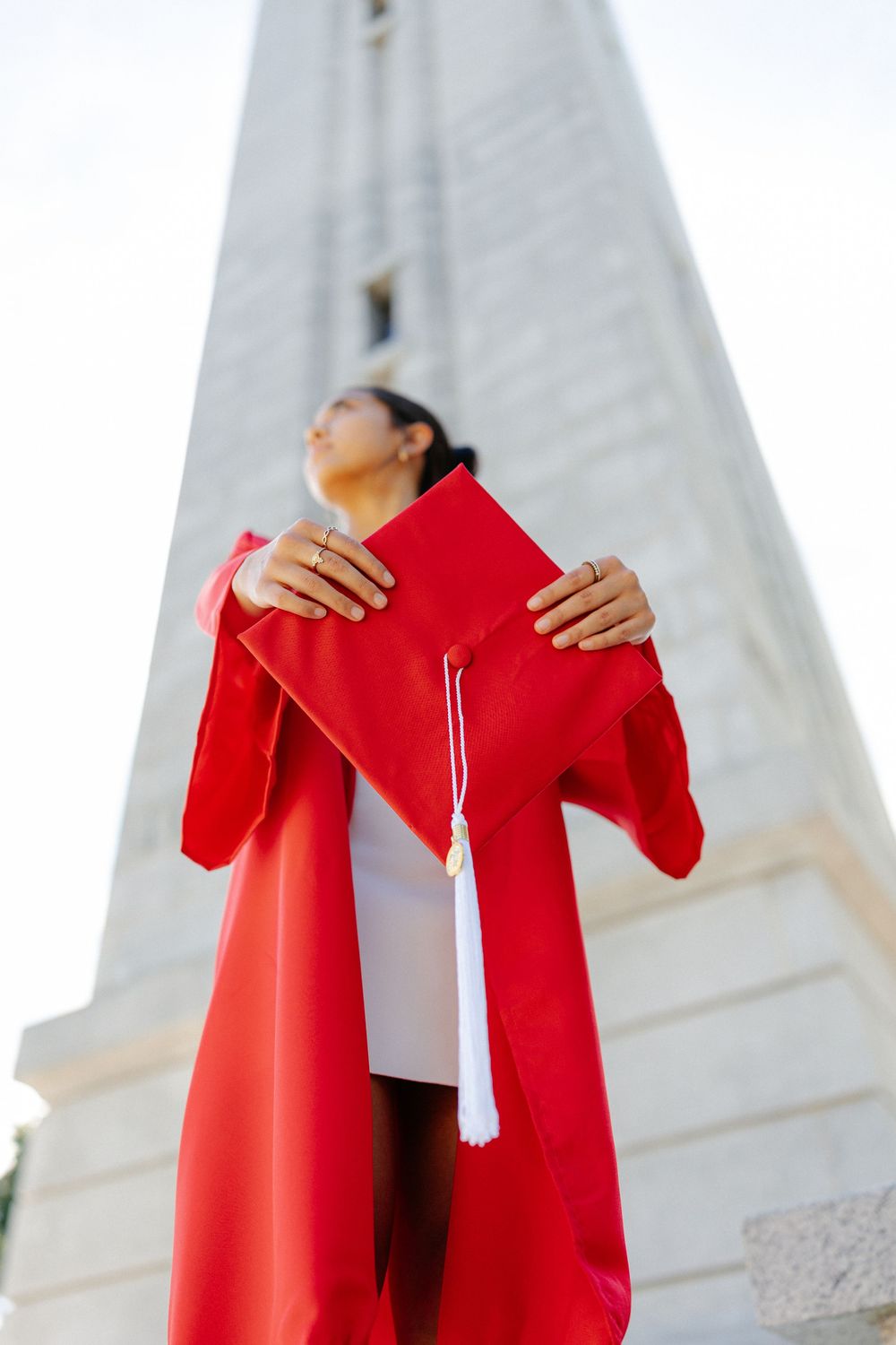 NC State Graduate in red gown holds cap up near tall monument against bright sky.
