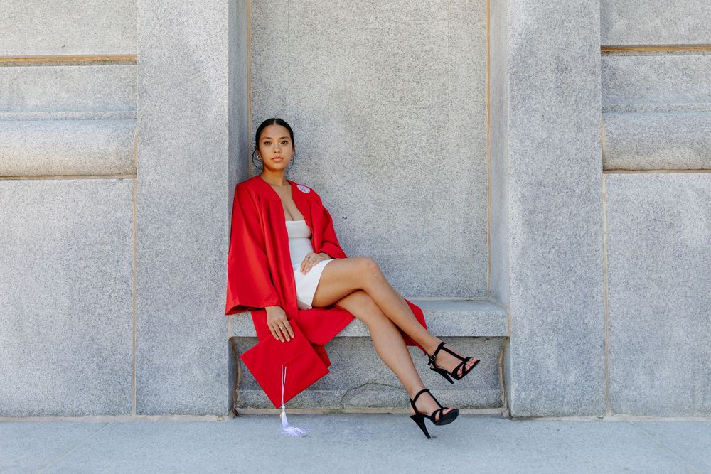 NC State Graduate in red graduation gown sits on stone bench against gray wall wearing heels.