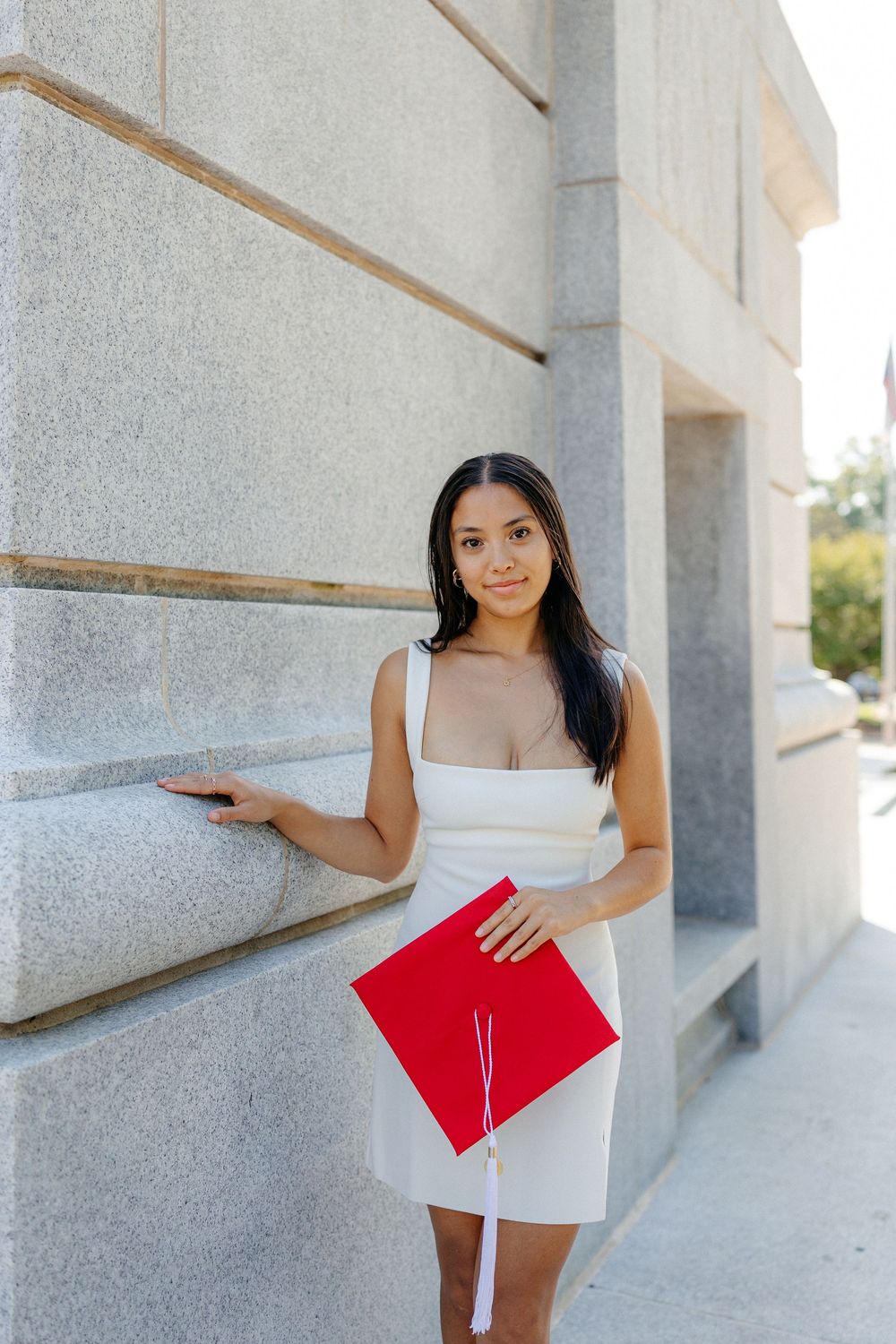 Young woman in white dress holds red graduation cap while leaning against stone wall.