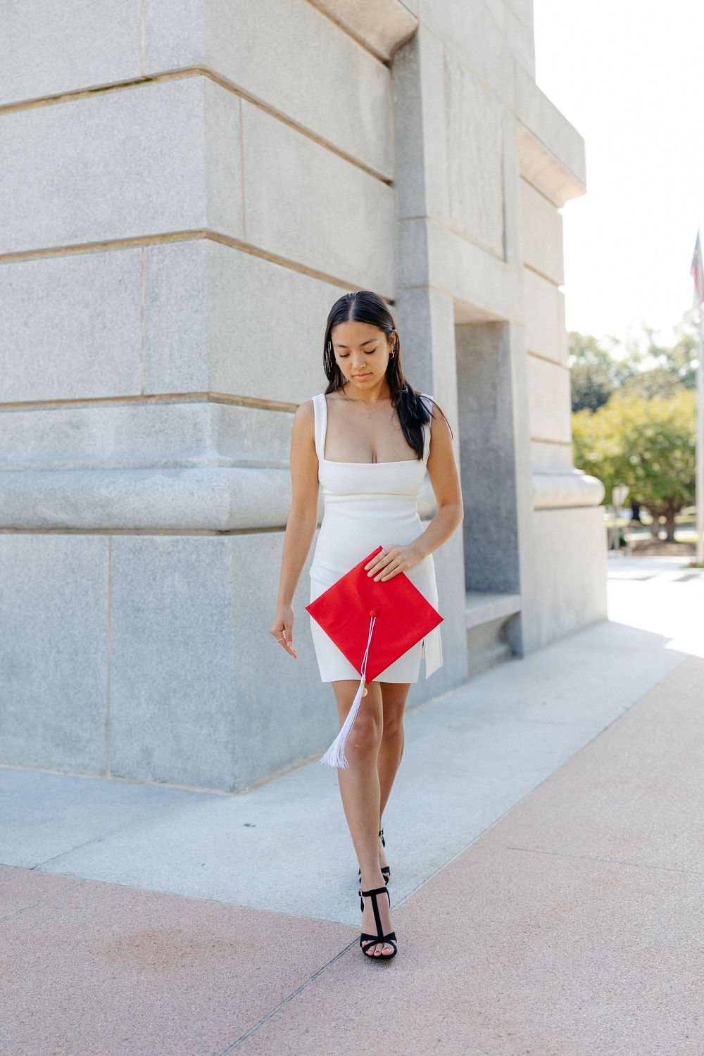 NC State Graduate in white dress carries red graduation cap while walking along stone building exterior.