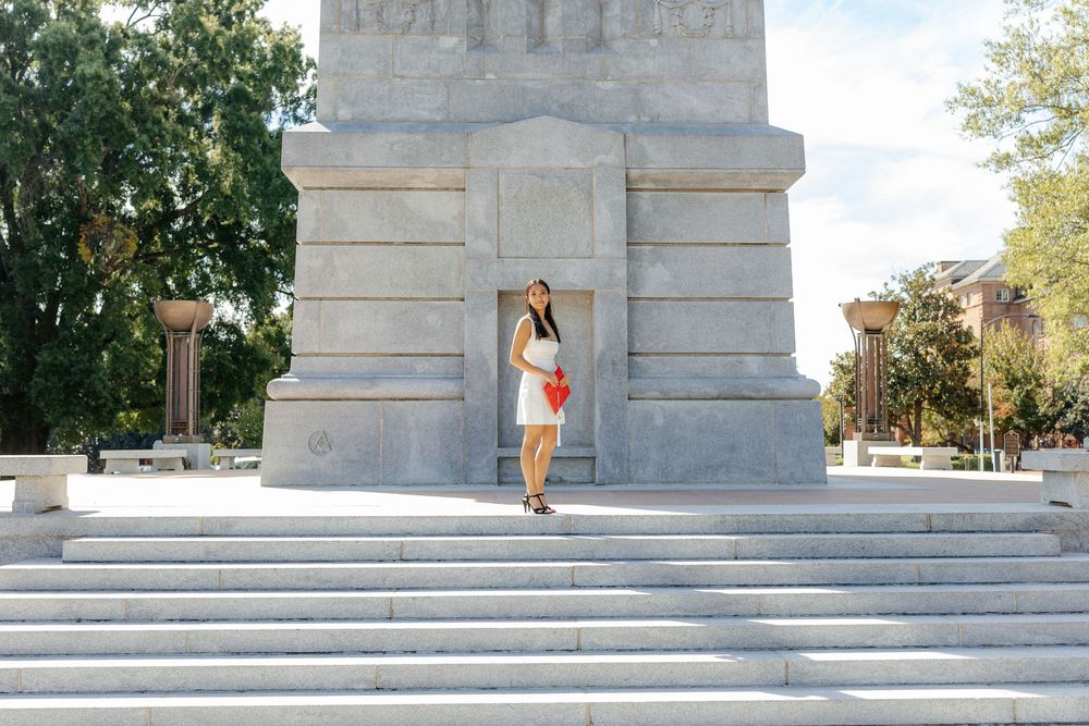 NC State Graduate in white dress holds red graduation cap while standing on steps of stone monument.