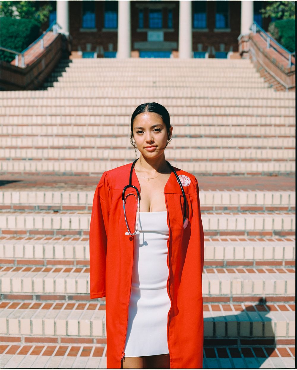NC State Graduate wearing red gown and stethoscope poses on brick steps, celebrating nursing school graduation.