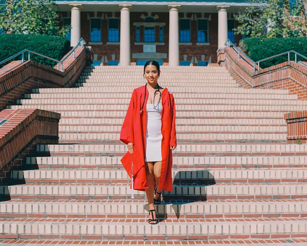 NC State Graduate in red gown and white dress stands on wide brick staircase in front of columned university building.