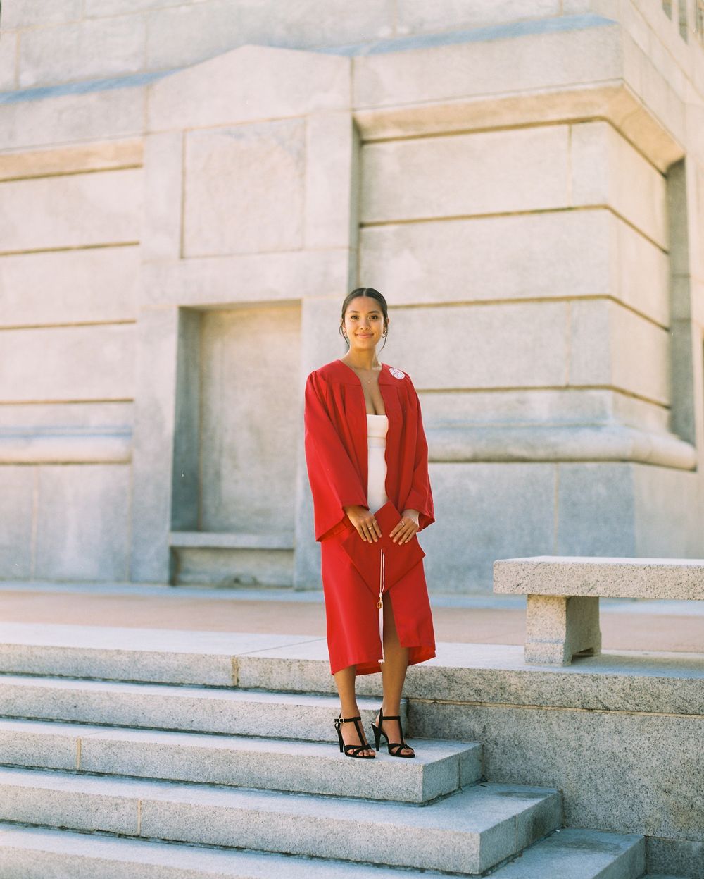 NC State Graduate in red academic gown stands on stone steps against classical stone wall on campus.