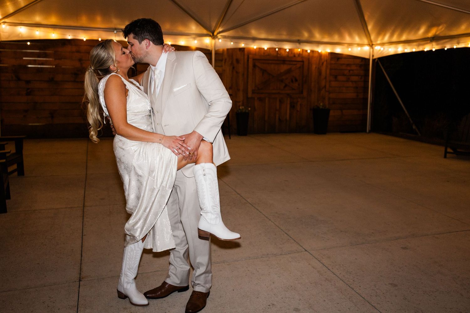 Bride and groom share romantic first dance kiss in white wedding attire under a tent with warm string lights.