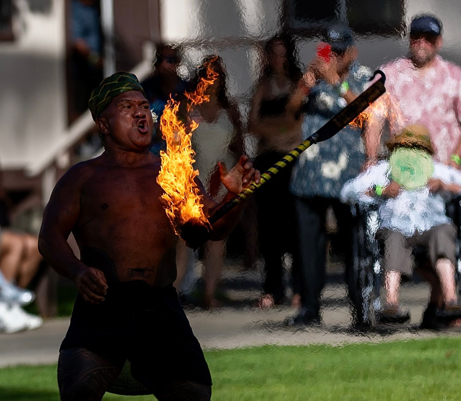 A shirtless fire dancer performs with flaming batons outdoors at a Hawaiian cultural event with spectators watching.