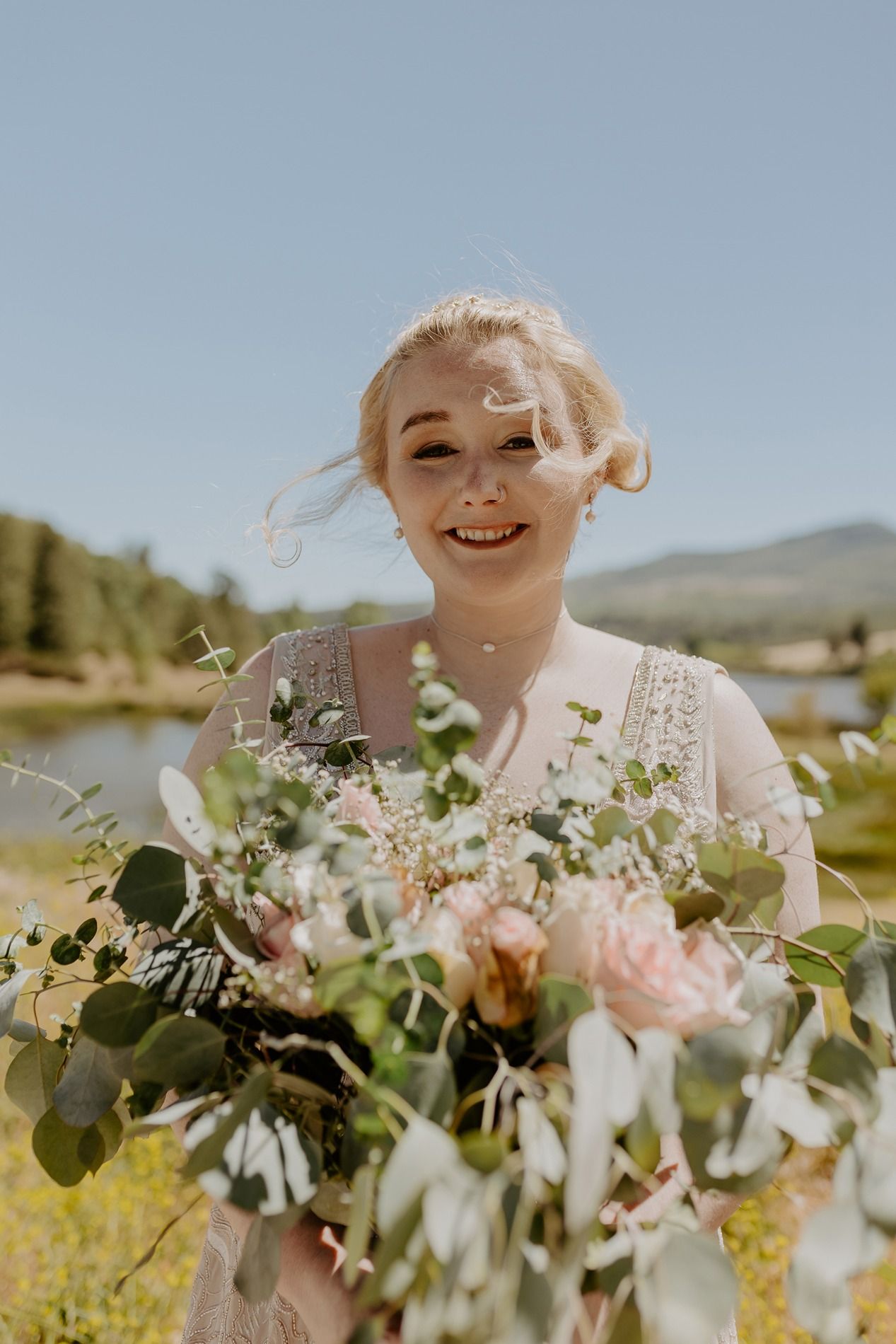Smiling blonde bride holding a large bouquet of pink roses and eucalyptus outdoors on a sunny day at Lake Cuyamaca, Julian.