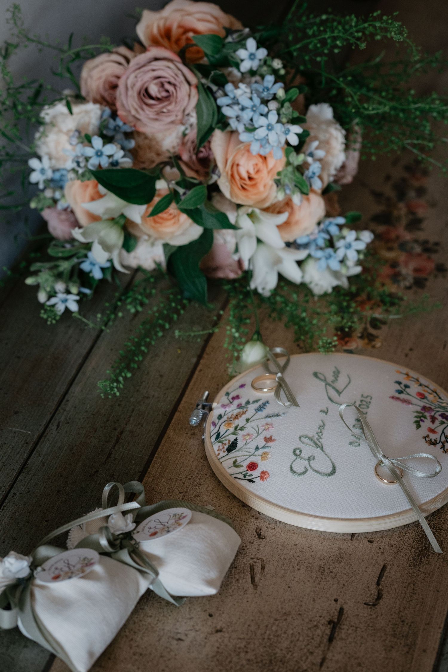 Wedding flat lay with peach roses, blue flowers, embroidered ring dish, and linen ring pillow on rustic wood table.