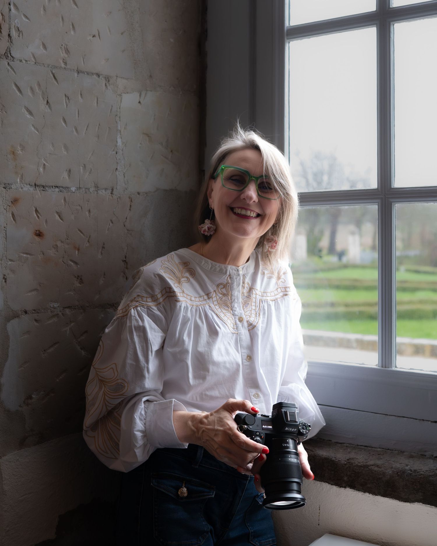 La souriante photographe Stéphanie Lecaudé tenant un appareil photo près d'une fenêtre d'un domaine d'exception