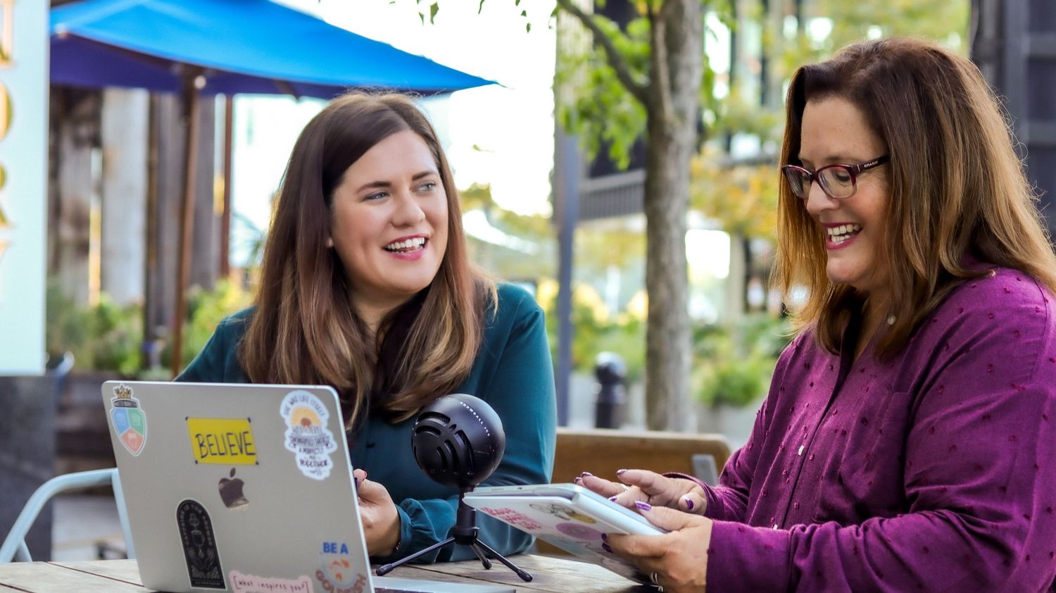 Two women smiling and collaborating outdoors at a cafe table with a laptop and microphone on a sunny day.