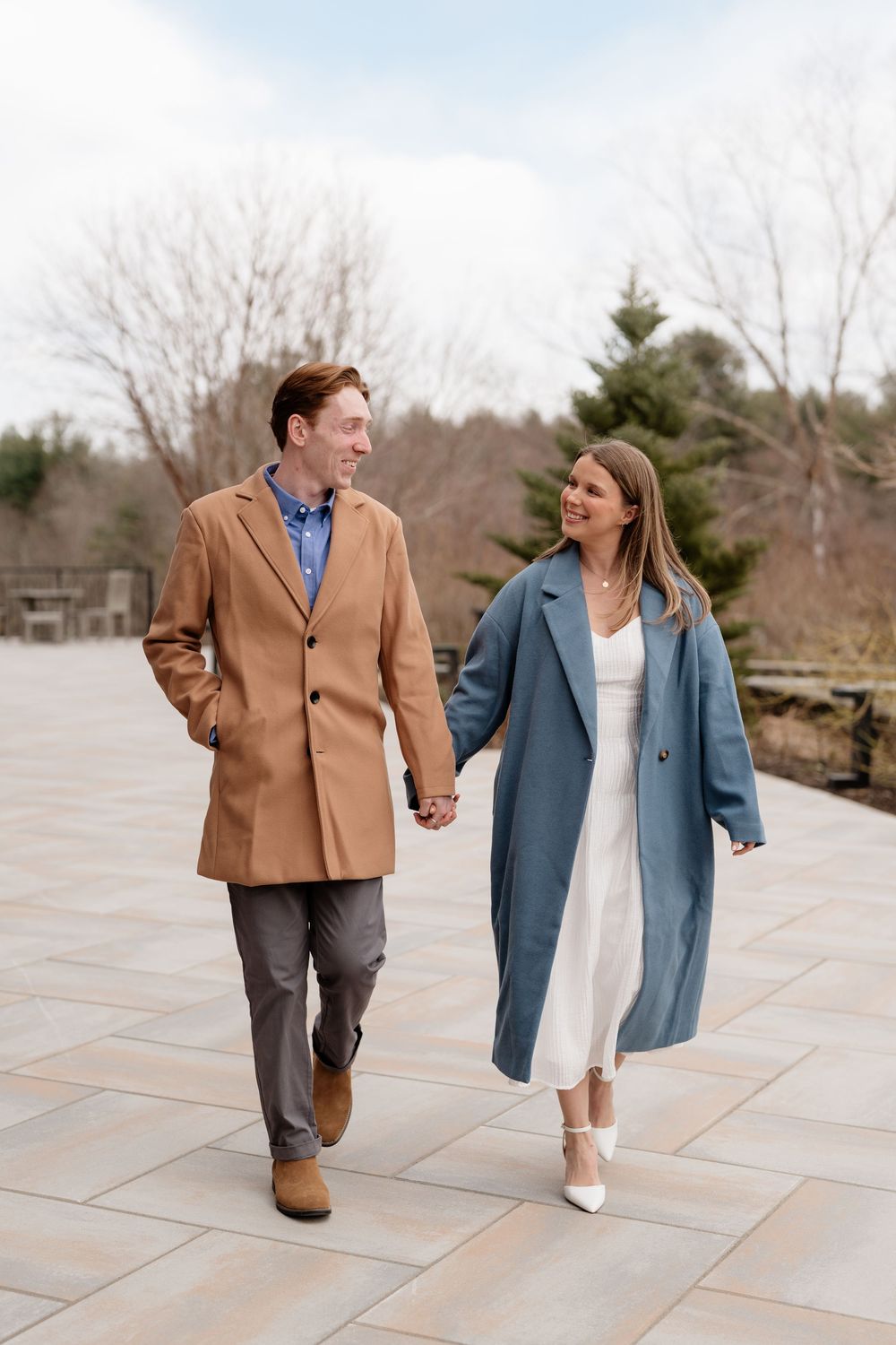 A couple walks hand-in-hand outdoors, man in tan coat, woman in blue coat and white dress, smiling at each other.