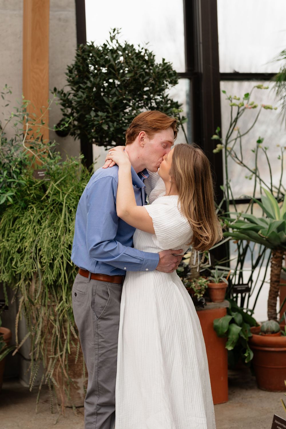 Couple sharing a tender kiss in a lush indoor greenhouse, man in blue shirt embracing woman in white dress.