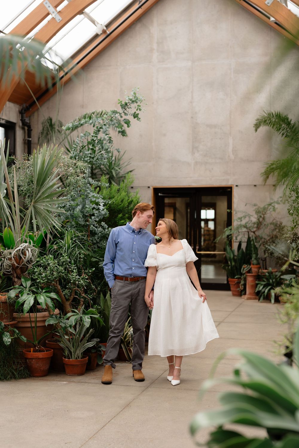 Couple walking hand in hand in a lush greenhouse conservatory, woman in white dress, man in blue shirt.