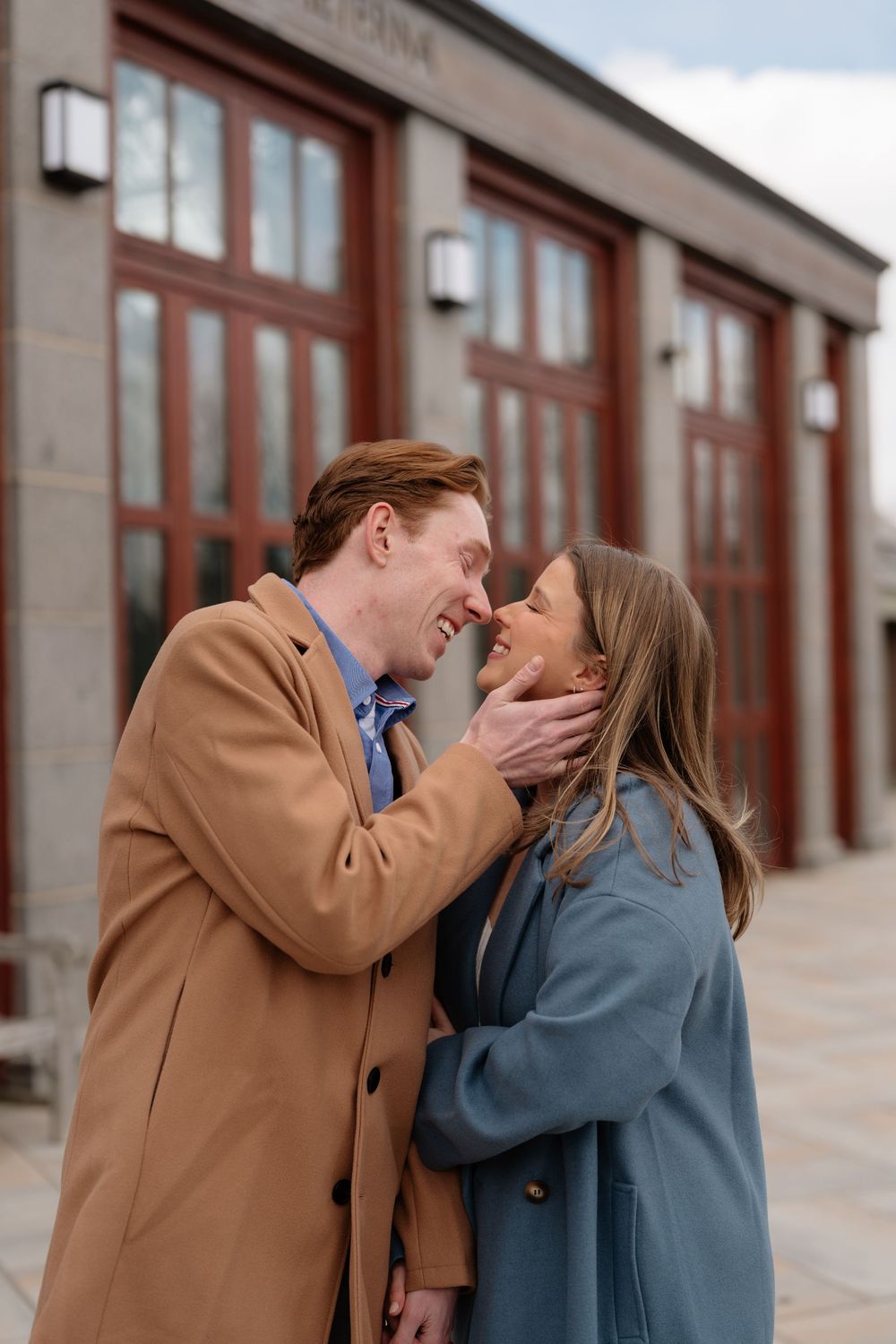 Couple sharing a romantic moment outdoors, man in tan coat gently holding womans face, both smiling and about to kiss.