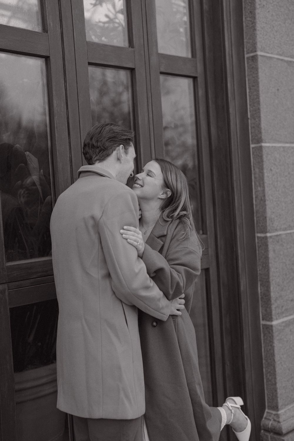Black and white photo of a romantic couple embracing and kissing outside a building with large windows.