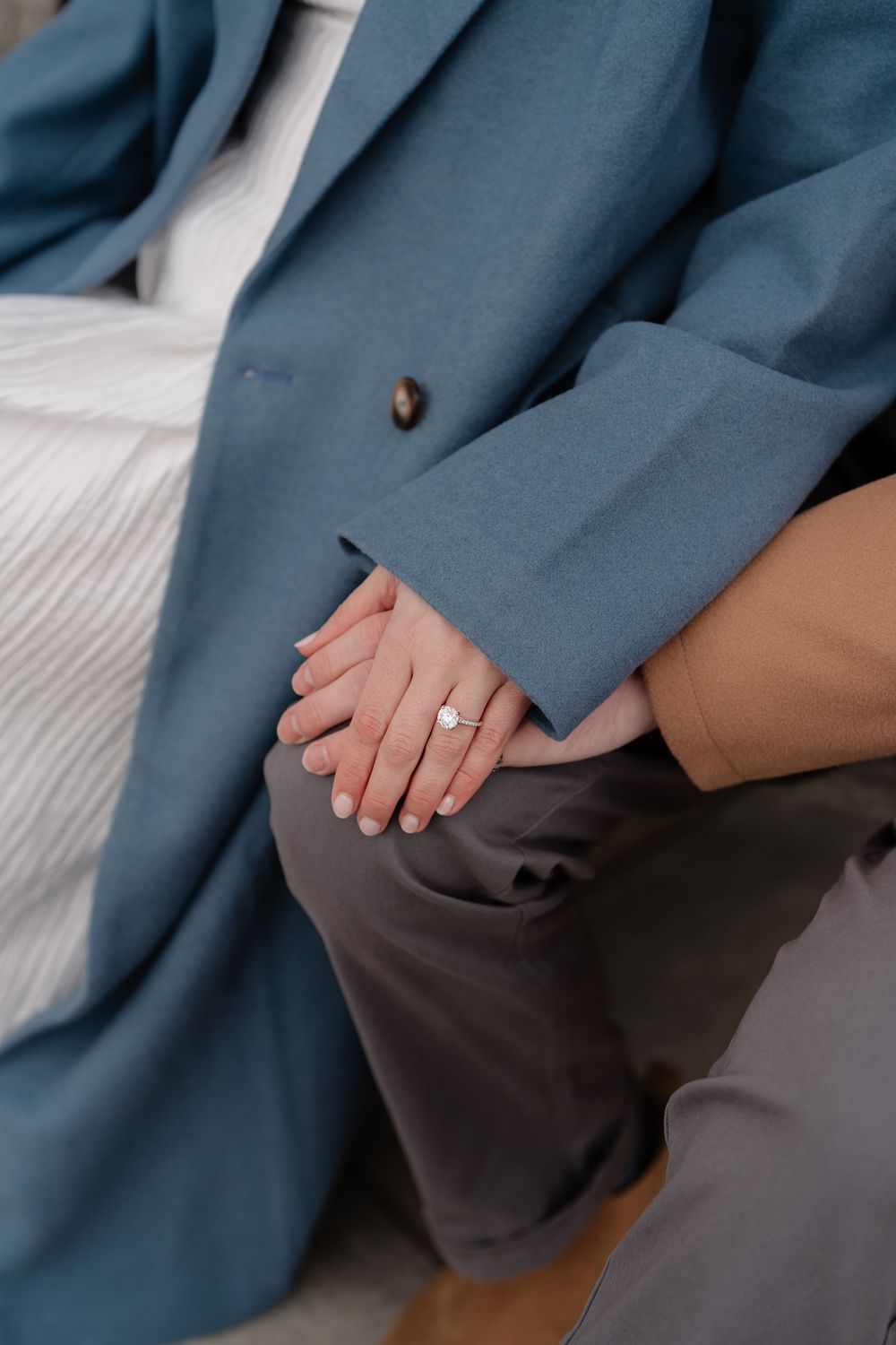 Close-up of couple holding hands with engagement ring, wearing blue blazer and tan coat while seated.