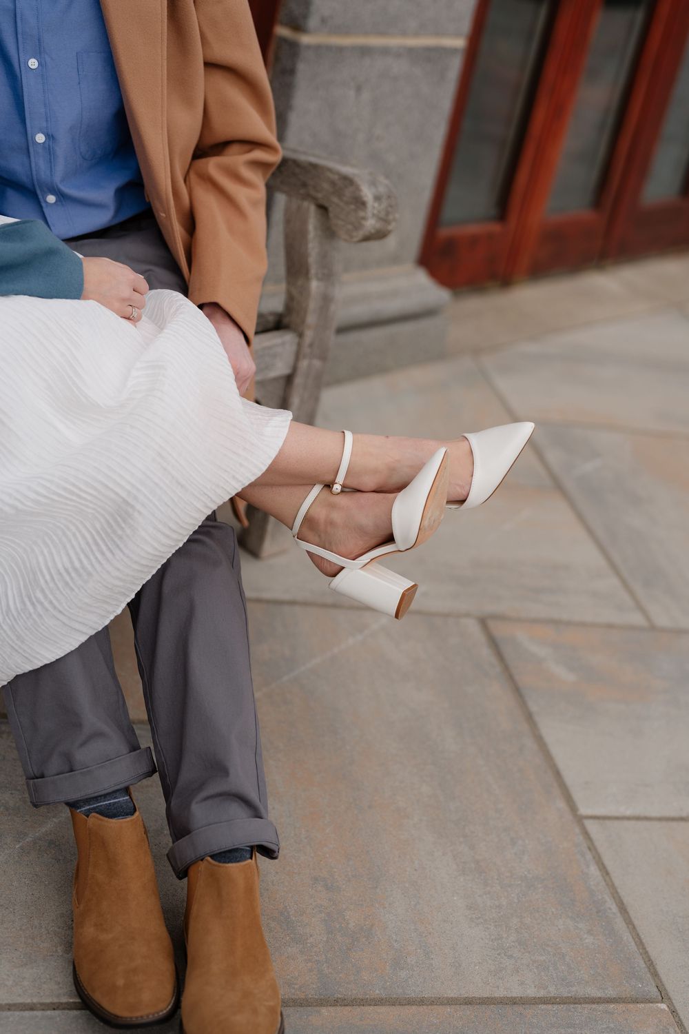 Couple sitting on a bench, woman in white dress and heels, man in gray pants and brown suede boots on stone patio.