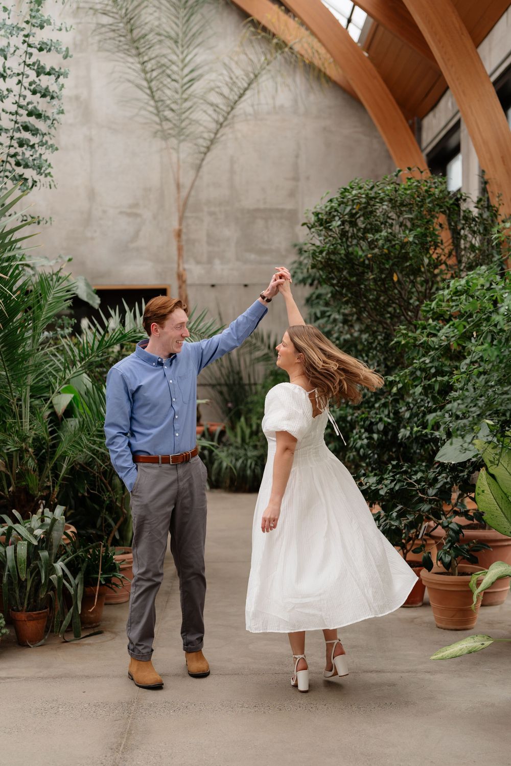 A couple dances in a lush greenhouse, man in blue shirt twirling woman in white midi dress among tropical plants.