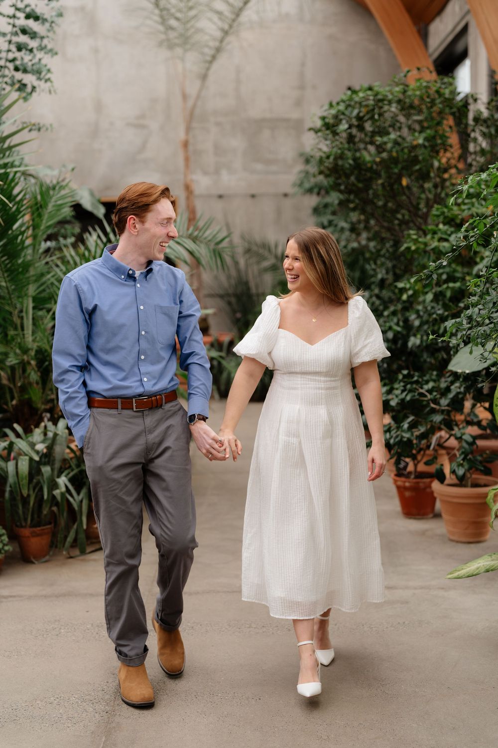A couple holds hands and smiles while walking through a greenhouse, man in blue shirt and woman in white dress.