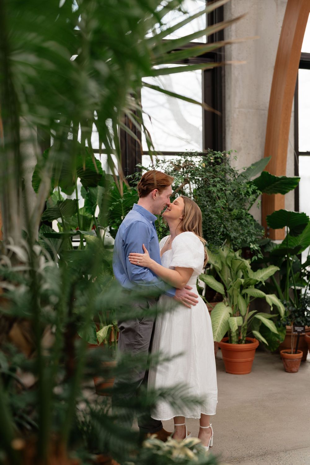 Romantic couple sharing a kiss surrounded by lush tropical plants in a greenhouse setting.