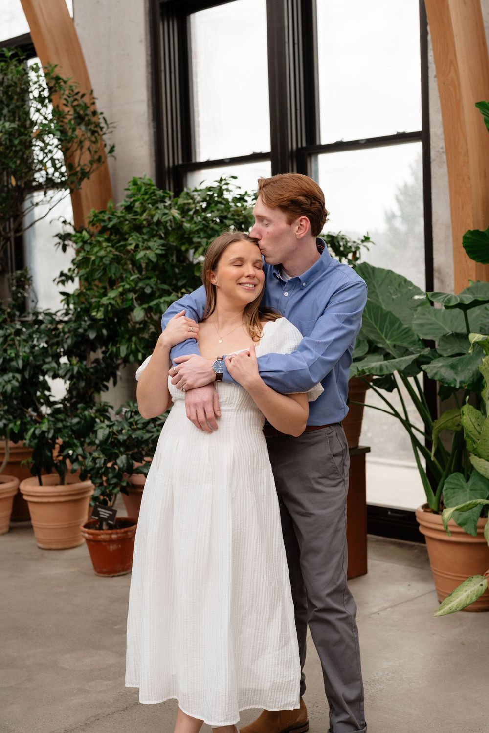 Couple embracing in a greenhouse, man kissing woman's temple, she wears a white dress surrounded by lush potted plants.