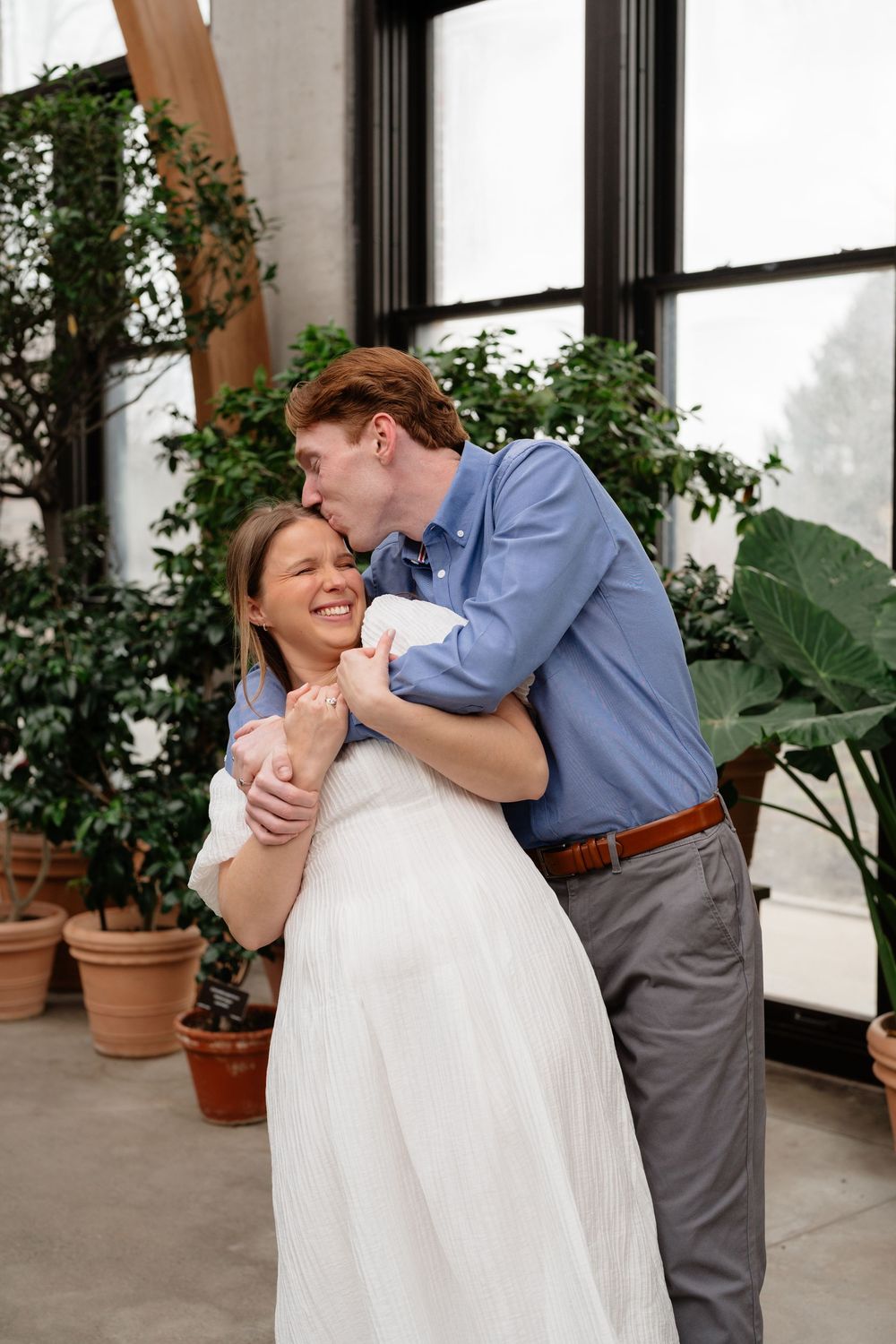 A man in a blue shirt kisses a smiling pregnant woman in a white dress in a sunlit greenhouse.