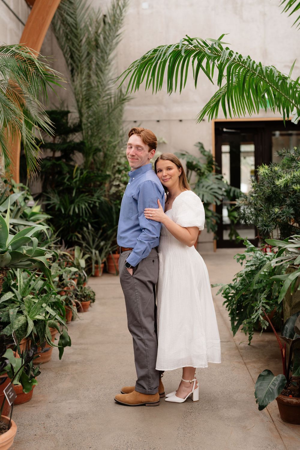 Couple posing together in a lush indoor botanical garden, woman in white dress embracing man in blue shirt.