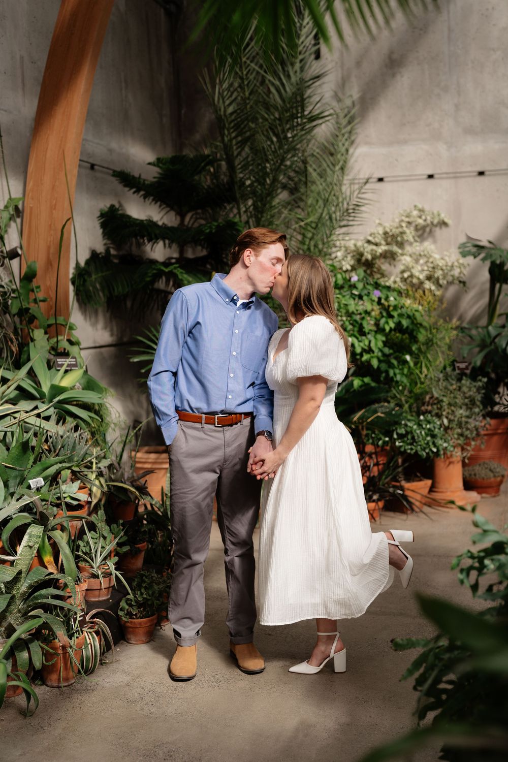 Couple kissing in a lush greenhouse, woman in white puff-sleeve dress, man in blue shirt and gray pants.