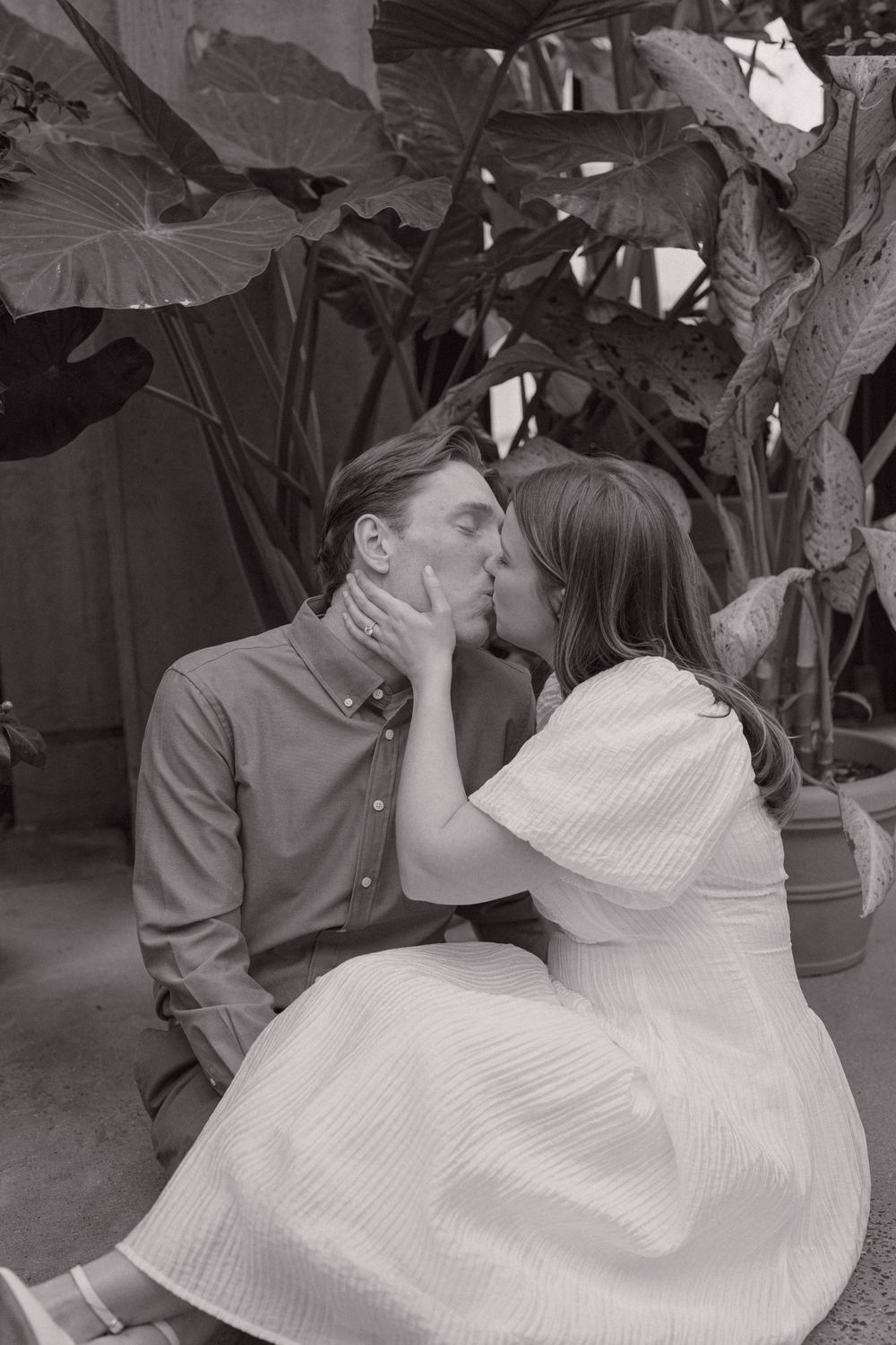 Black and white photo of a couple sharing a romantic kiss surrounded by tropical plants.
