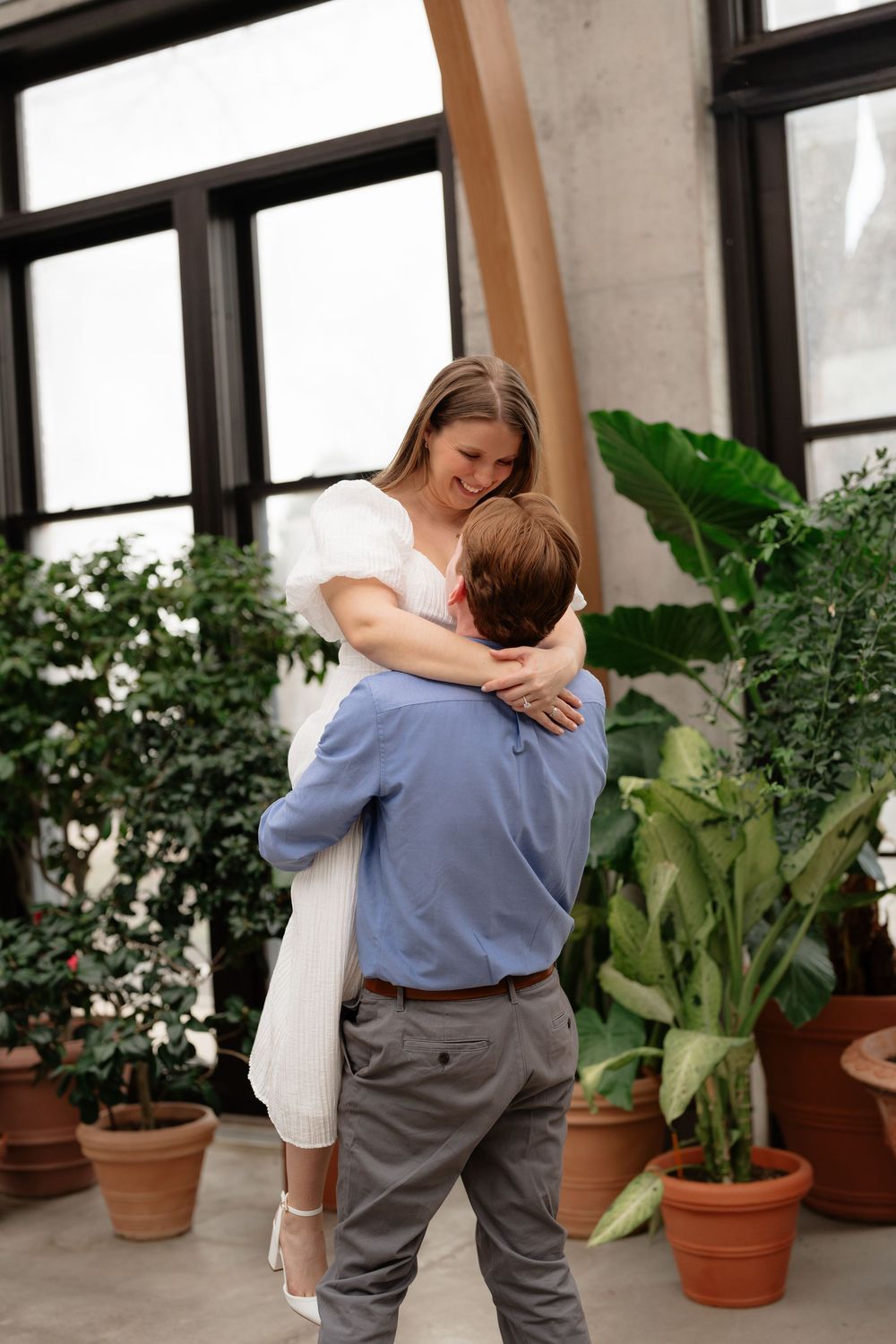 Happy couple sharing a romantic lift embrace in a sunlit greenhouse filled with tropical potted plants.