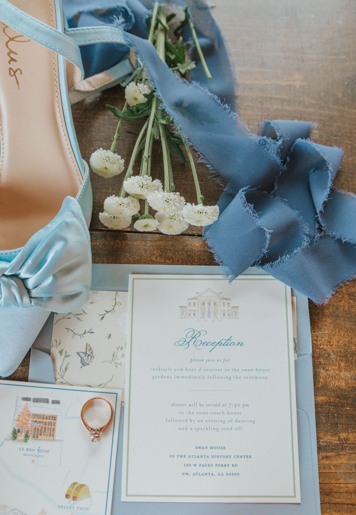 Dusty blue wedding flat lay with heels, white ranunculus, silk ribbon, reception invitation, and rose gold ring on wood.