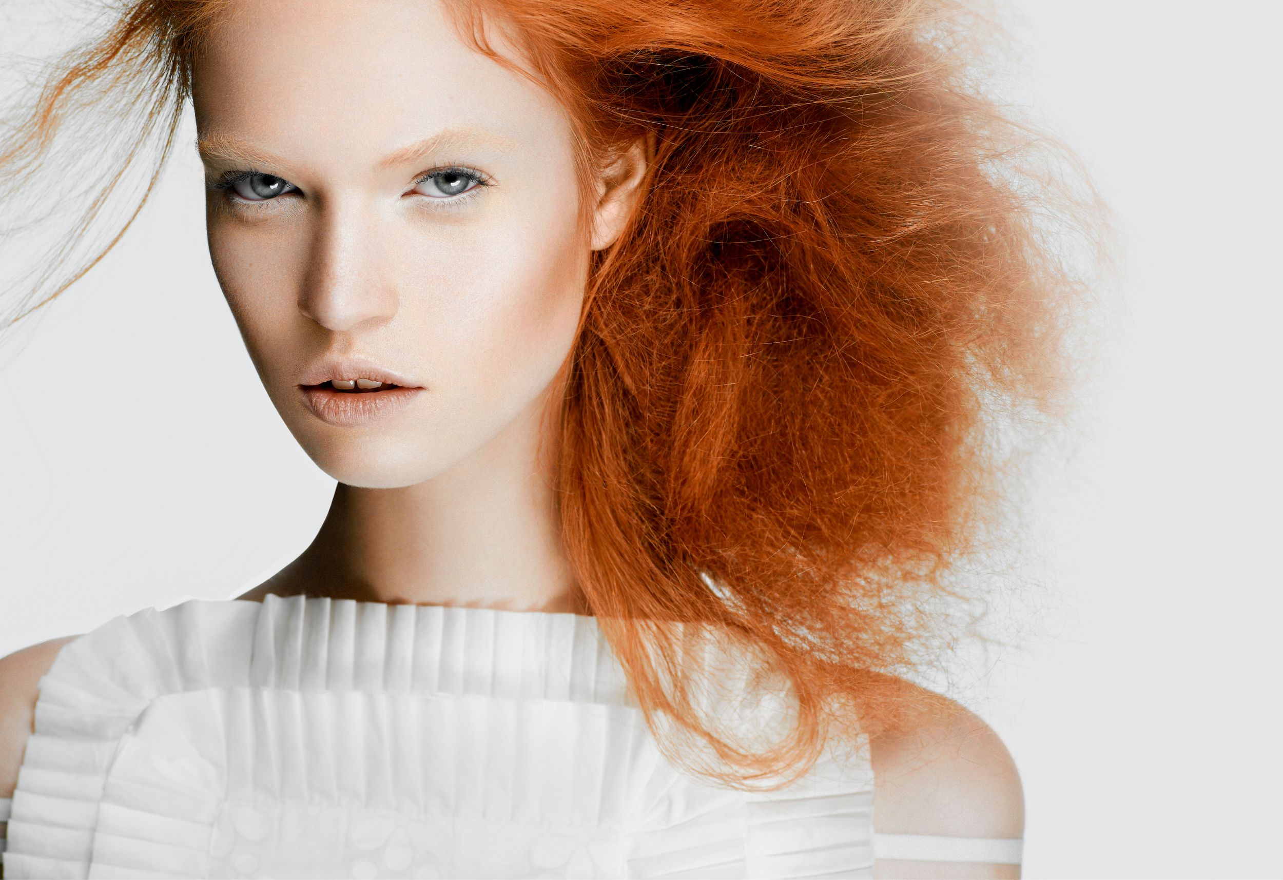 A striking model with voluminous red hair wearing a white structured top against a light background.