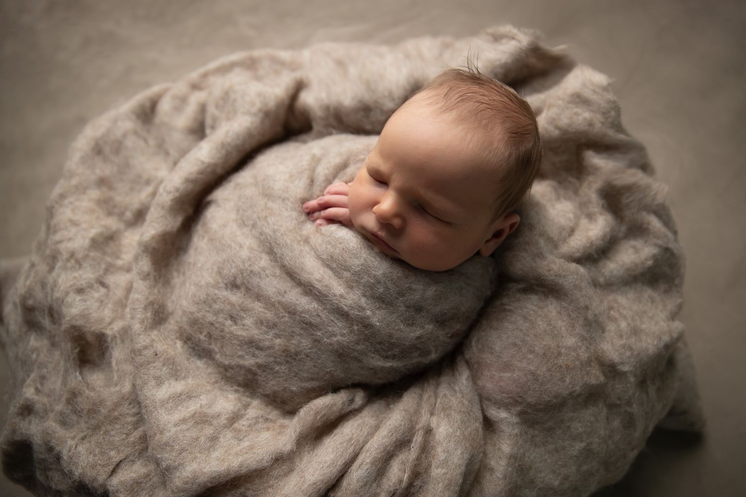 Peacefully sleeping newborn baby wrapped snugly in a soft gray knit blanket on a neutral backdrop.