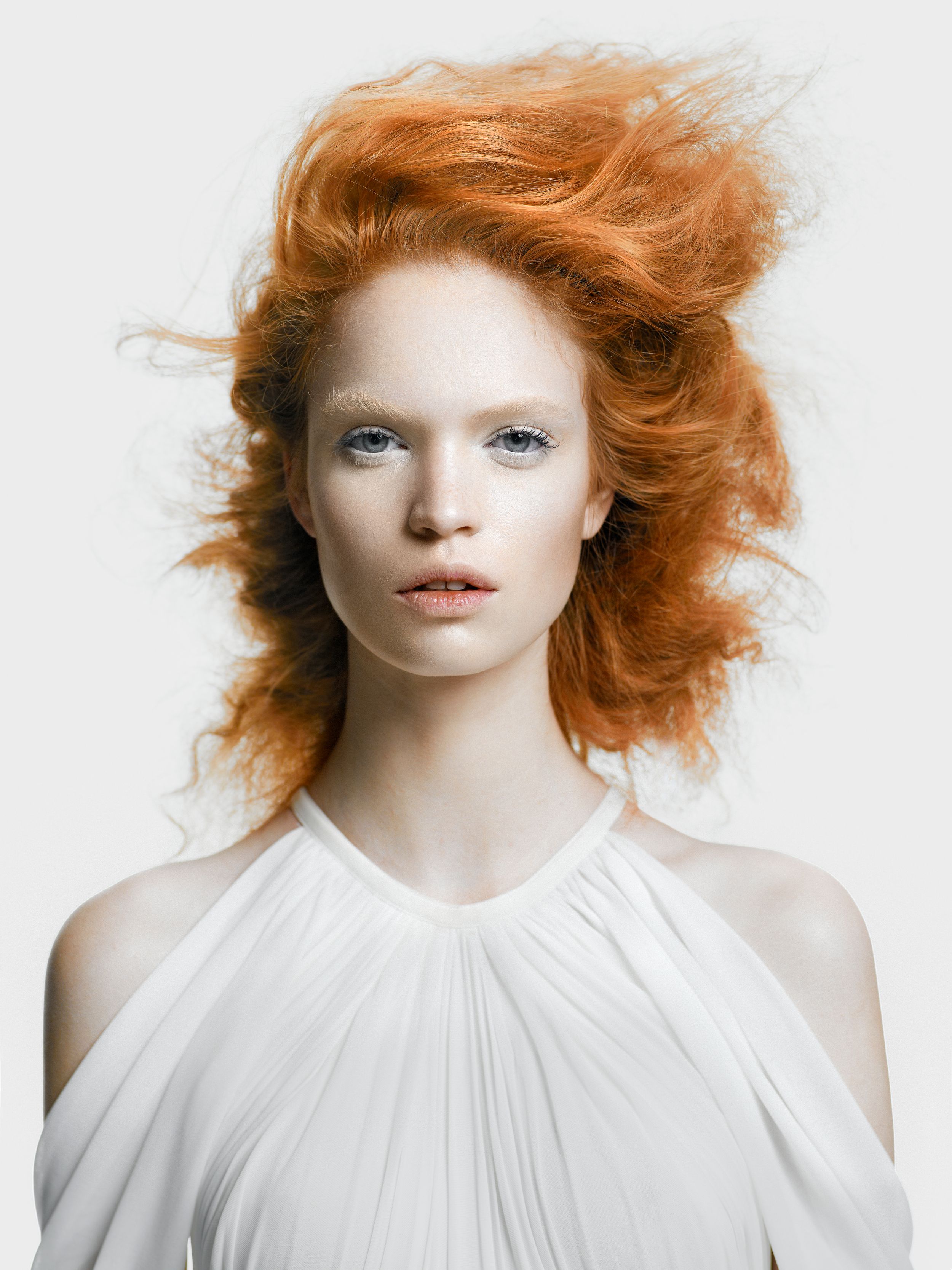 Portrait of a pale woman with voluminous red hair wearing a white halter dress against a light background.