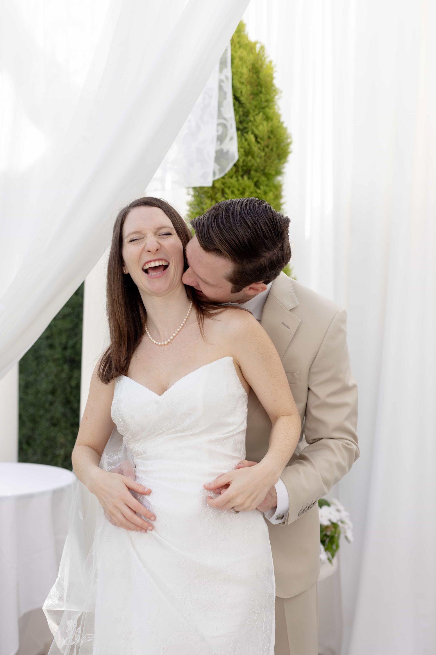 Groom in tan suit kissing laughing bride on cheek under white draped wedding ceremony canopy outdoors.