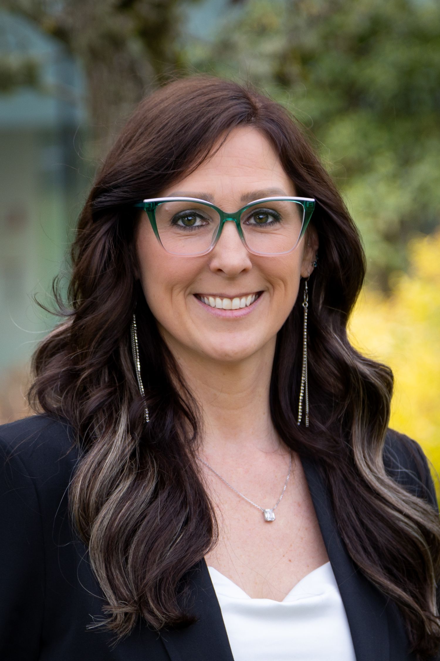 Professional woman with green glasses and dark hair smiling outdoors in a black blazer and white top.