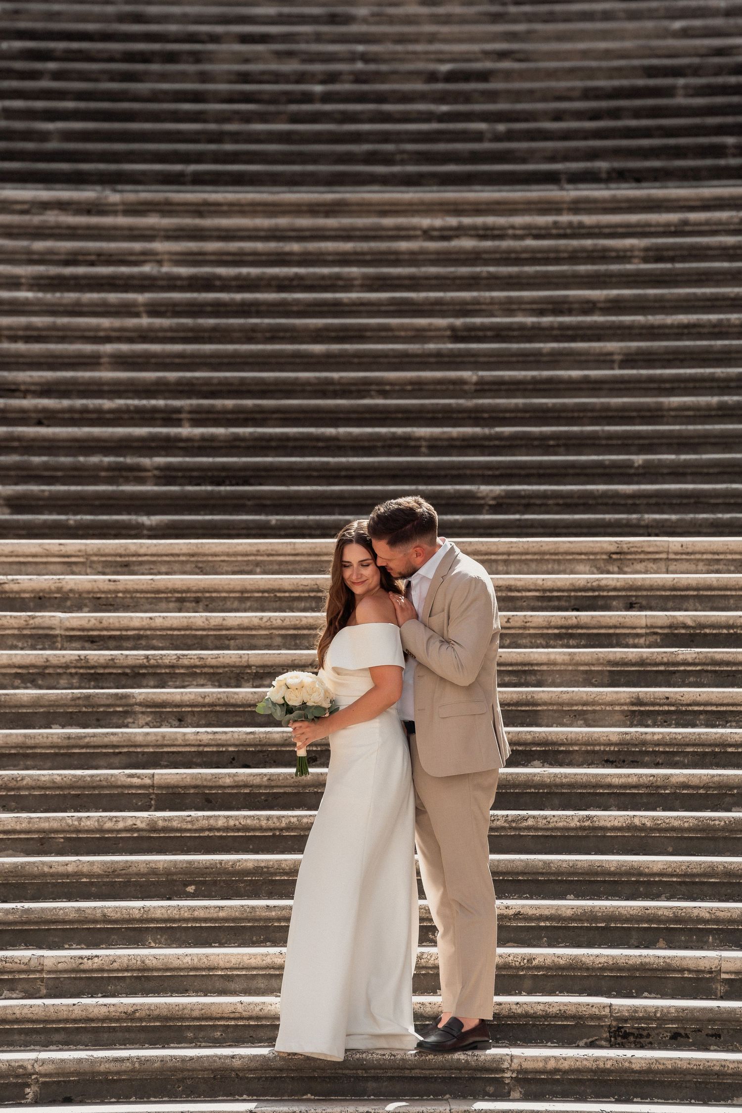 Bride and groom embracing on grand stone steps, bride in white off-shoulder gown holding bouquet, groom in beige suit.
