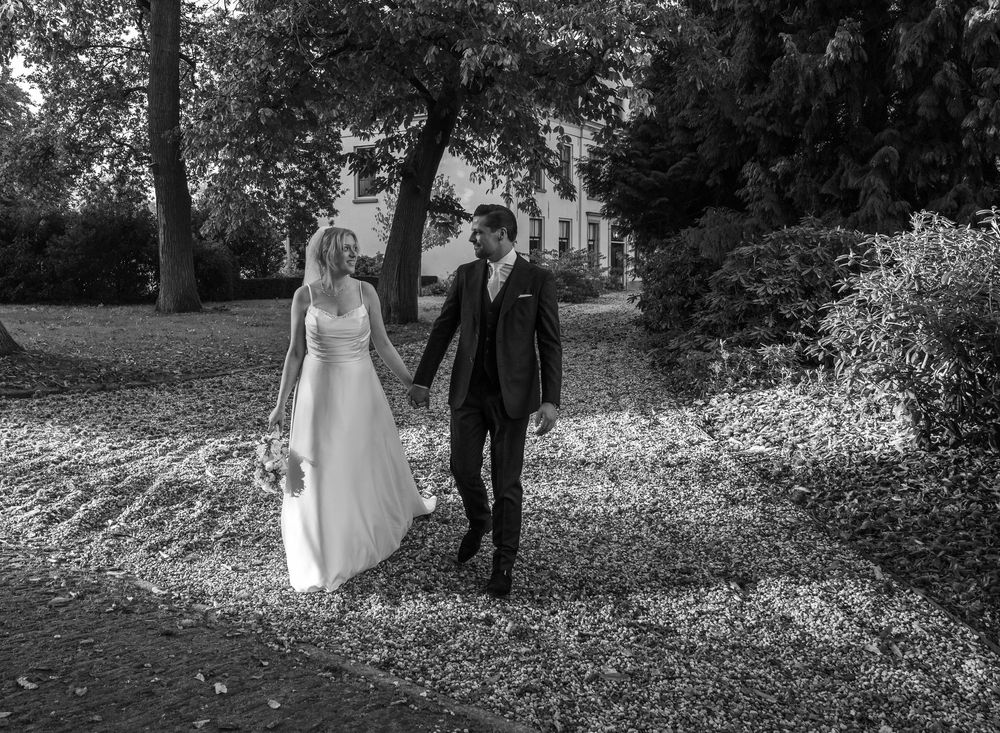Zwart-witfoto van een pasgetrouwd stel dat hand in hand loopt over een zonovergoten, met bomen omzoomd tuinpad. Trouwfotograaf t harde