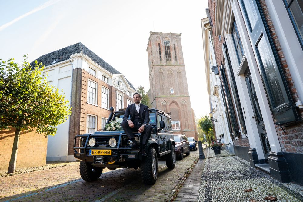 Bruidegom in donker pak zit op een zwarte Land Rover op een geplaveide Nederlandse straat met een kerktoren op de achtergrond. In elbrug