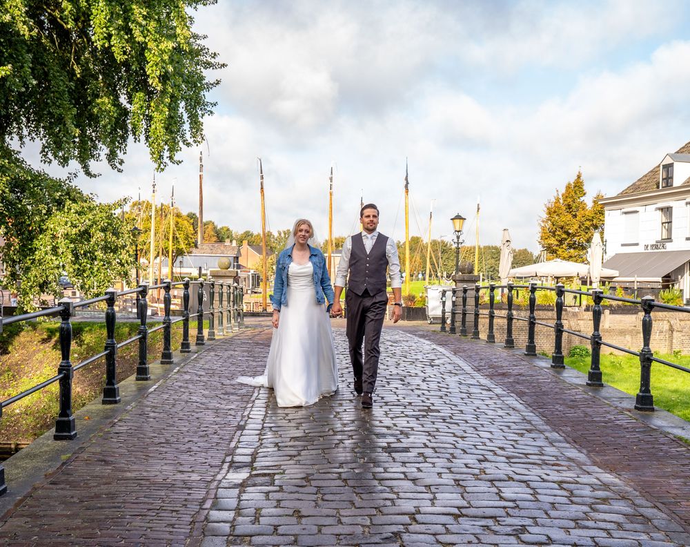 Bruid en bruidegom lopen hand in hand over een geplaveide brug in een Nederlandse haven op hun trouwdag. In Elburg trouwfotograaf