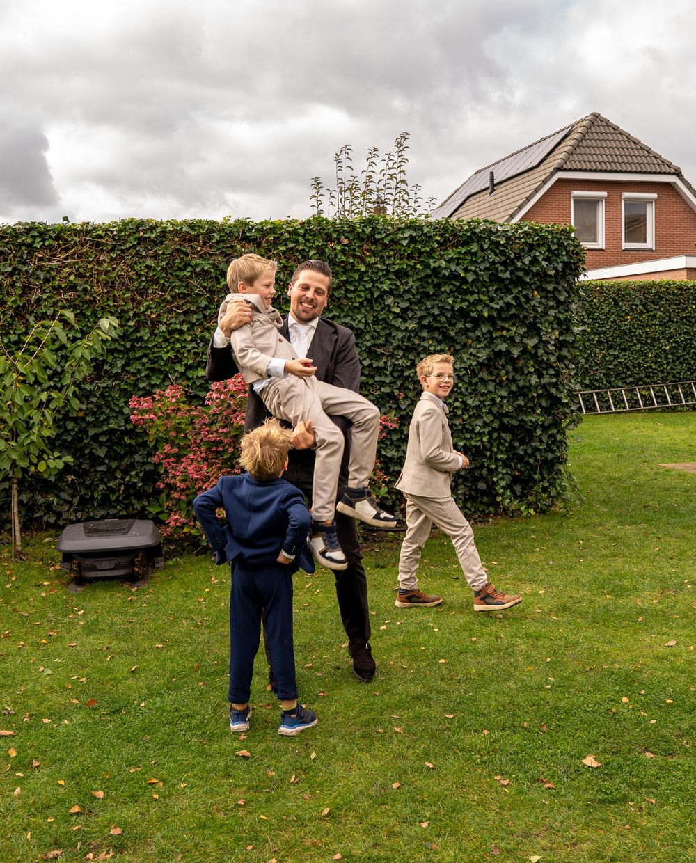 Een opgewekte bruidegom in trouwkleding tilt zijn kind op terwijl hij met drie kinderen in een groene achtertuin speelt.