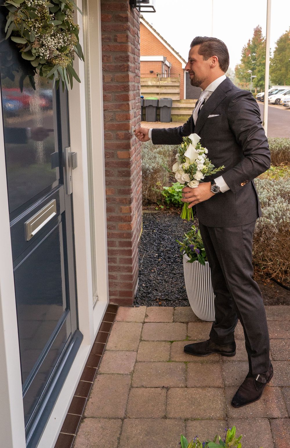 Een elegante bruidegom in een donker pak houdt een boeket witte bloemen vast terwijl hij aanbelt bij een bakstenen huis.