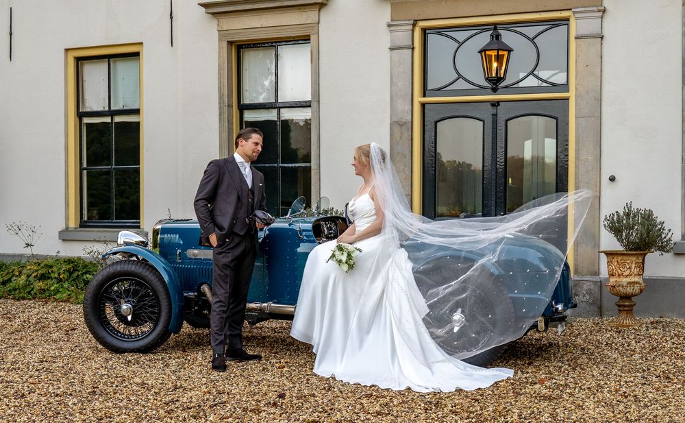 Bruid en bruidegom poseren naast een vintage blauwe klassieke auto voor een elegant landhuis op hun trouwdag.