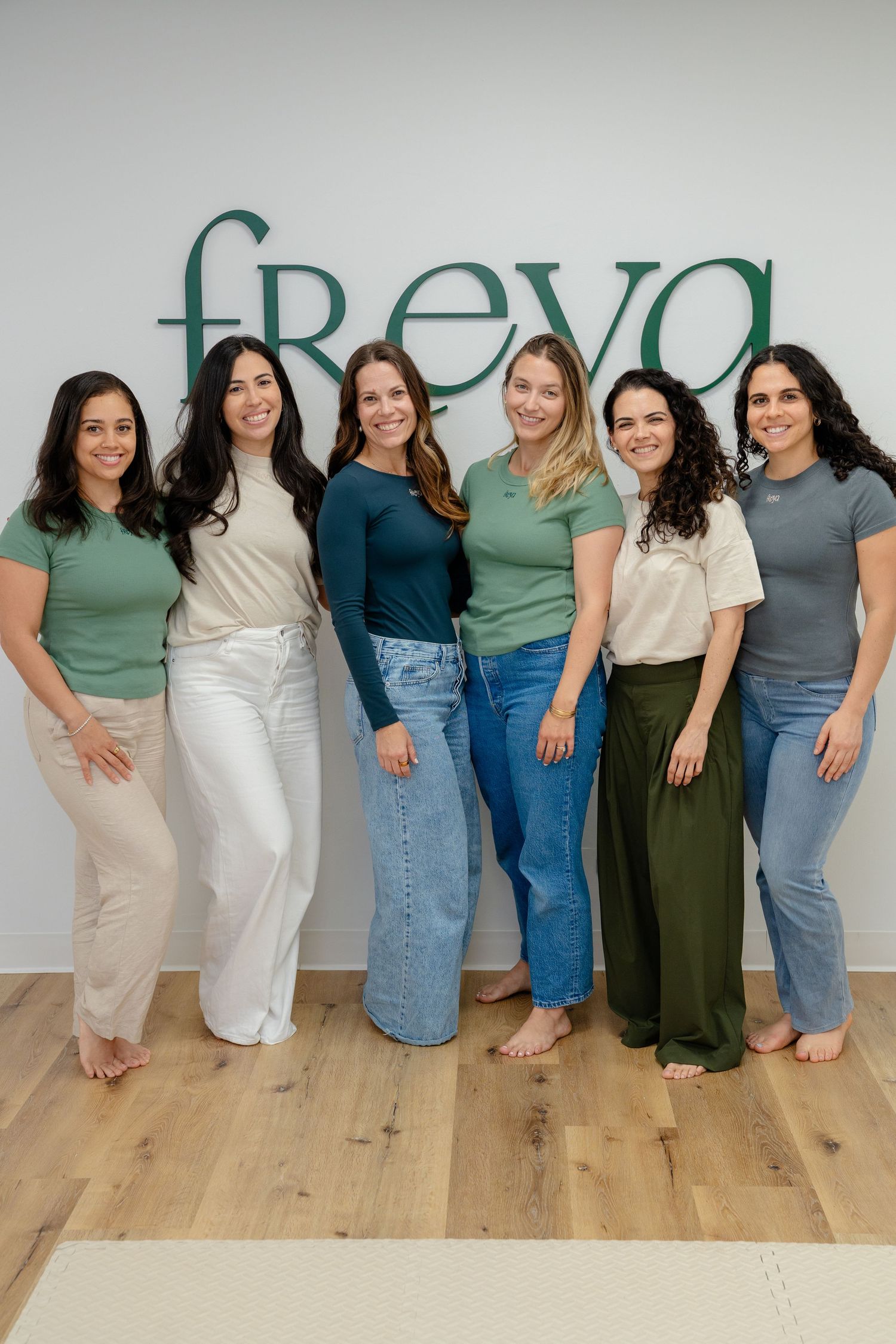 Six women in green and neutral tones pose smiling in front of a Freya branded wall on a wood floor.