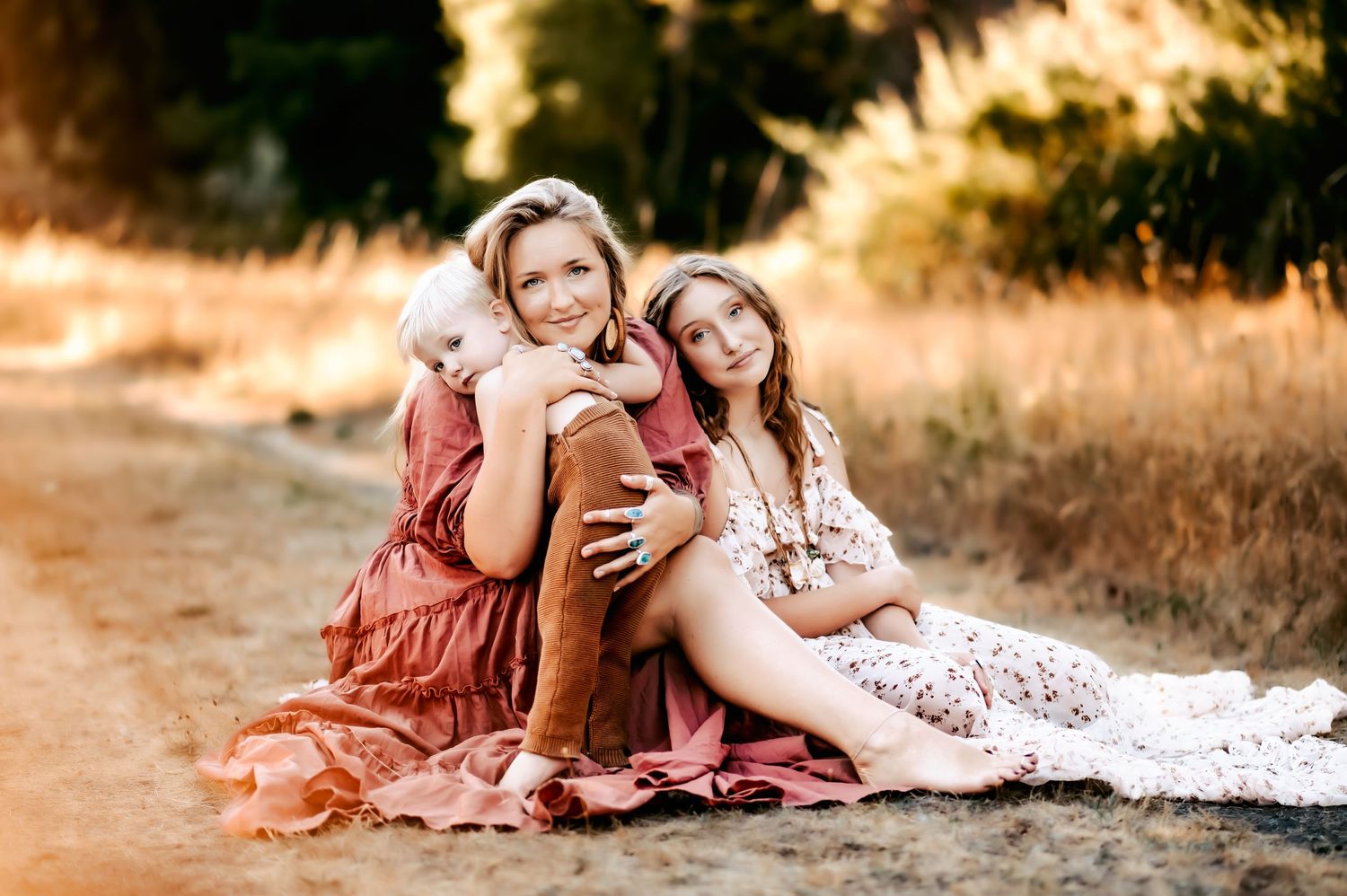 A mother and two daughters share a warm embrace on a sunlit dirt path surrounded by golden fields.