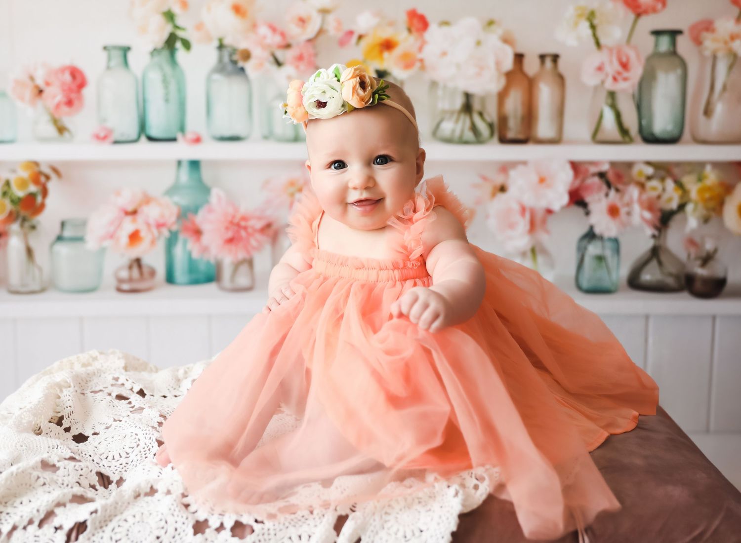 Smiling baby girl in peach tulle dress with floral headband, sitting before colorful flower vases backdrop.