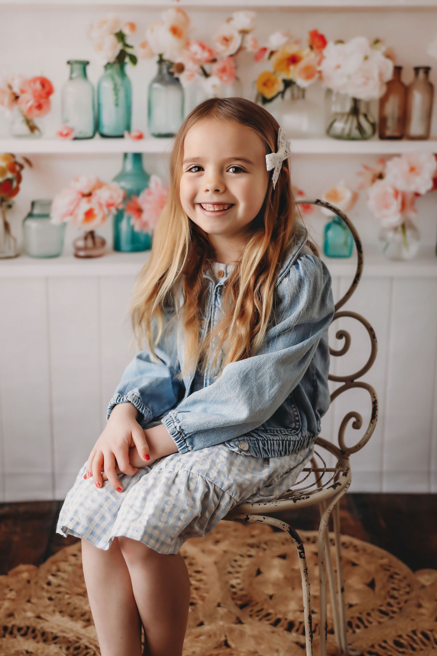 Smiling girl with long blonde hair in denim jacket and gingham dress sits on ornate chair with floral decor.
