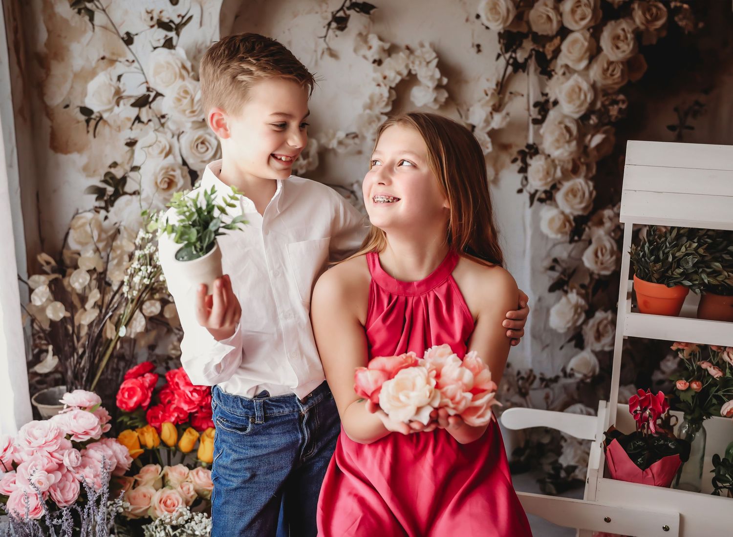 A boy and girl in a flower-filled studio, smiling and holding bouquets, dressed in white shirt and red dress.