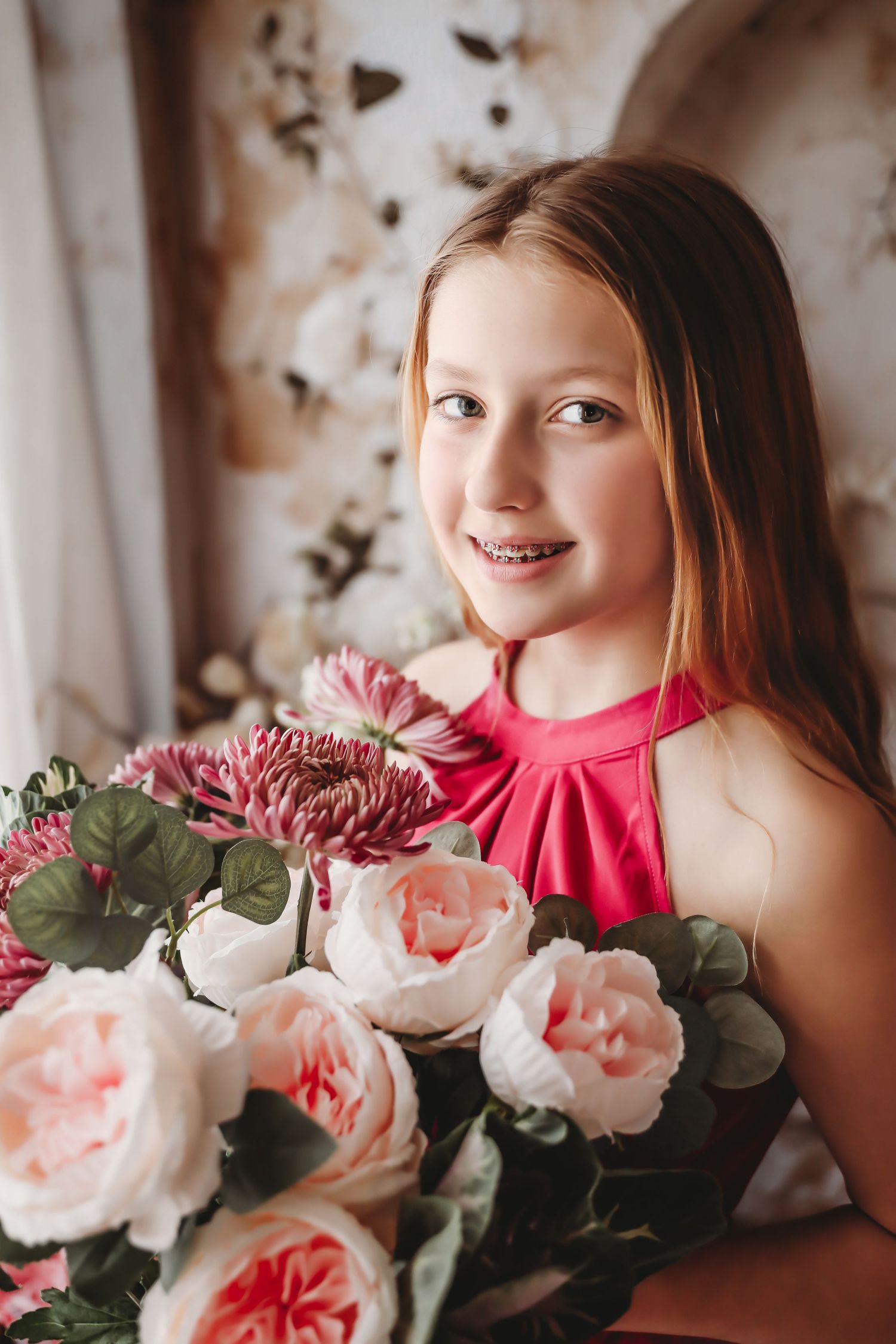 Smiling young girl with braces holding a large bouquet of pink roses and chrysanthemums in a floral studio setting.