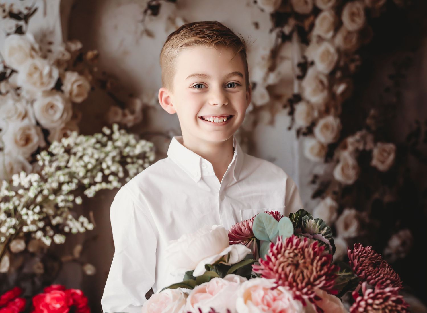 Smiling boy in white shirt holding a bouquet of pink and red flowers with a floral backdrop.
